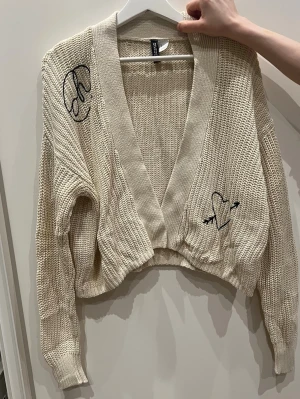 Beige cropped stickad kofta H&M - Trendig beige stickad kofta från H&M med croppad passform och djup V-ringning. Koftan har svarta broderade detaljer, bland annat ett hjärta med pilar och en grafisk bokstav på axeln. Långa ärmar och bred ribbad kant nedtill. Perfekt till lager på lager.