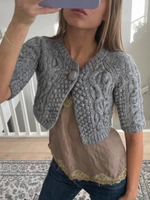 Grå stickad bolero med flätmönster - Kort grå stickad bolero med halvlånga ärmar och snyggt flätmönster. Stängs med en matchande stickad knapp framtill. Perfekt att slänga över en topp för en trendig lager-på-lager-look. Mjuk och skön i toppen skick 💕