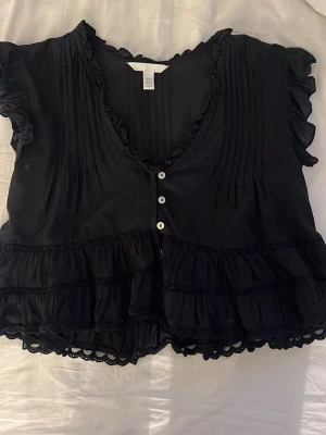 Svart volangblus med knappar H&M - Söt svart blus från H&M med volangdetaljer, v-ringning och korta ärmar. Blusen har dekorativa knappar framtill, rynkade detaljer och spetskant längst ner. Croppad modell som ger en trendig look.
