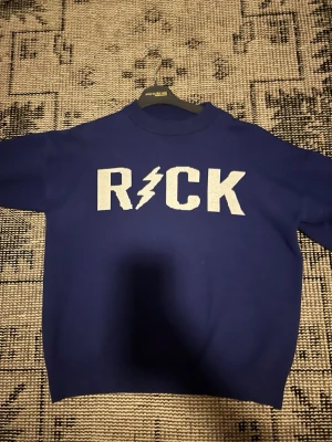 Blå sweatshirt med ROCK-tryck - Cool blå sweatshirt från Mini Villains med stort vitt 'ROCK'-tryck och blixt på bröstet. Tröjan har rund halsringning och ribbade muddar. Perfekt för dig som gillar en edgy och avslappnad stil.