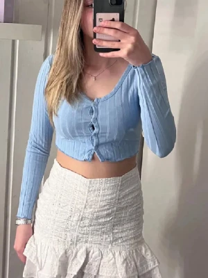 Topp -  ljusblå croppad topp med lång ärm. Toppen har ribbad struktur och knappar framtill 