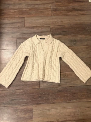 Beige kabelstickad tröja BikBok - Säljer en beige kabelstickad tröja från BikBok med krage och v-ringning. Tröjan har breda ärmar och ribbade muddar vid nederkant. Perfekt för dig som gillar en avslappnad men trendig look. Tröjan är endast använd två gånger. 