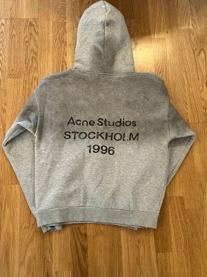 Grå hoodie från Acne Studios - Snygg grå hoodie från Acne Studios med tryck på ryggen där det står 'Acne Studios STOCKHOLM 1996'. Klassisk passform med huva och ribbade muddar vid ärmslut och nederkant. Perfekt för en avslappnad och trendig look.