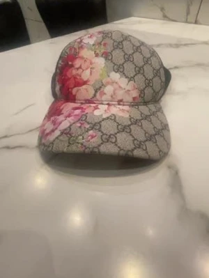 Gucci keps med blommigt mönster - Snygg keps från Gucci med klassiskt GG-mönster i grått och svart, dekorerad med rosa och röda blommor framtill. Kepsen har böjd skärm och justerbar baksida. Perfekt accessoar för dig som vill sticka ut.