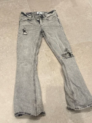 Grå slitna bootcut jeans Gina Tricot - Säljer ett par grå bootcut jeans från Gina Tricot med slitningar på låret och knät. Jeansen har fem fickor, hög midja och en snygg tvättad look. Perfekta för dig som gillar en avslappnad och trendig stil.