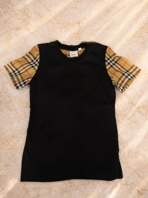 T-shirt med rutiga ärmar från Luxury - Unik t-shirt med klassiska rutiga ärmar i beige, svart och rött. Finns i både svart och vitt med kontrasterande kropp. Perfekt för dig som vill sticka ut med en trendig och ungdomlig stil. Tillverkad i mjuk bomull för skön känsla hela dagen.