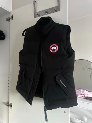 Svart dunväst från Canada Goose - Snygg svart dunväst från Canada Goose. Västen har hög krage, dragkedja och knappar framtill samt praktiska fickor. Perfekt för lager-på-lager och håller dig varm under kyliga dagar. Väldigt fin i storlek S och skulle kunna passa en M. 