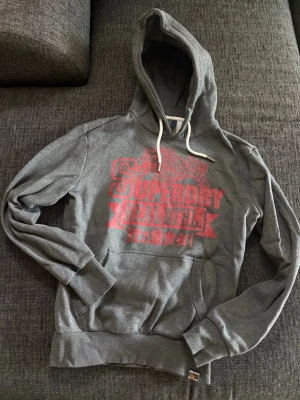 Grå hoodie från Superdry - Säljer en mörkgrå hoodie från Superdry med röd logotyp. Hoodien har vita tjocka snören i huvan.