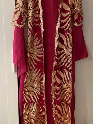 Kurdisk newroz kaftan klänning - Unik röd långklänning med transparenta långärmar och iögonfallande broderade gulddetaljer i ett organiskt mönster. Klänningen har öppen front och skimrande detaljer som verkligen sticker ut. Perfekt för dig som vill ha något extra speciellt.