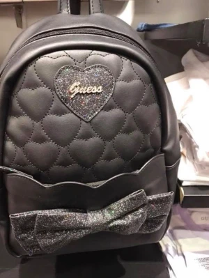 Svart ryggsäck med glitterdetaljer Guess som NY - Snygg svart ryggsäck från Guess i konstläder med quiltade hjärtan, glittrande hjärtformad logga och stor glittrig rosett framtill. Justerbara axelband och praktiskt fack. Perfekt för dig som vill ha en trendig och söt accessoar. Som NY
