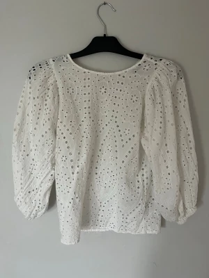 Vit broderad blus med puffärm - Säljer en vit blus med broderade hålmönster och puffiga trekvartsärmar. Blusen har rund halsringning och är gjord i ett lätt, luftigt tyg som känns som bomull. Perfekt för dig som gillar romantisk och trendig stil.