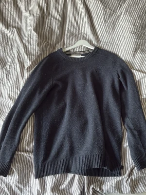 Mörkblå stickad tröja - En klassisk mörkblå stickad tröja med rund halsringning och ribbade muddar vid ärmslut och nederkant. Tröjan har en enkel och stilren design, perfekt för lager-på-lager och passar till det mesta. Materialet ser ut att vara ullblandning.