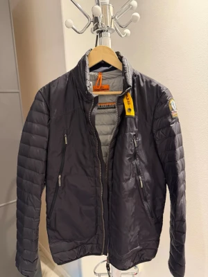 Grå pufferjacka från Parajumpers - Säljer en grå pufferjacka från Parajumpers med quiltade paneler och två dragkedjeförsedda fickor framtill. Jackan har en hög krage, gul detalj vid halsen och logopatch på ärmen. Perfekt för kalla vinterdagar och tillverkad i syntetmaterial.