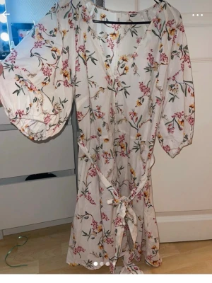 Vit blommig klänning med puffärm - Supersöt vit klänning med blommigt mönster i rosa och gult. Klänningen har puffiga trekvartsärmar, v-ringning och knytband i midjan för en snygg passform. Perfekt för varma dagar och somriga vibes!