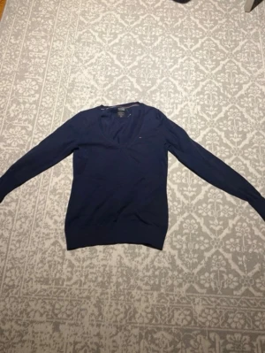 Mörkblå stickad tröja Tommy Hilfiger - Mörkblå stickad tröja från Tommy Hilfiger med v-ringning och lång ärm. Klassisk liten logga broderad på bröstet. Tröjan har ribbade muddar vid ärmslut och nederkant. Perfekt för lager-på-lager och enkel att matcha.