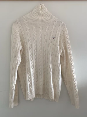 Vit kabelstickad polotröja Gant - Kabelstickad polotröja från Gant i mjuk vit bomull. Tröjan har hög polokrage, ribbade muddar och diskret Gant-logga broderad på bröstet. Klassisk och tidlös design som passar perfekt till kyligare dagar.
