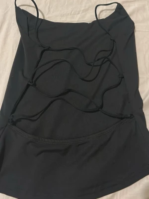 Svart linnetopp med snörning - Snygg svart linnetopp med tunna axelband och trendig snörningsdetalj framtill. Toppen har en tight passform och är perfekt för att skapa en edgy look. Tillverkad i mjukt syntetmaterial som känns skönt mot huden.