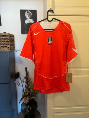 Röd Korea fotbollströja Nike 2004 reissue - Säljer en röd fotbollströja från Nike med Koreas landslagsmärke på bröstet. Tröjan har vita detaljer och mesh-paneler på ryggen för extra ventilation. Perfekt för träning eller match, snygg och sportig design.