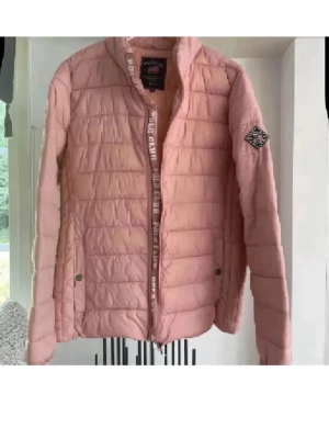Rosa pufferjacka från Polo Club - Ljusrosa pufferjacka från Polo Club med quiltad design och hög krage. Jackan har dragkedja framtill, två fickor med tryckknappar och en patch på ärmen. Snygg detalj med Polo Club-tryck längs dragkedjan. Perfekt för kalla vinterdagar.