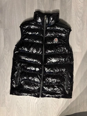 Svart dunväst från Moncler - Snygg svart dunväst från Moncler med glansigt material och hög krage. Västen har två dragkedjeförsedda fickor och Moncler-logga på bröstet. Perfekt för lager-på-lager och ger en clean streetwear-look.