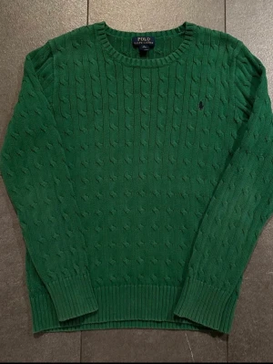 Grön kabelstickad tröja Polo Ralph Lauren - Snygg grön kabelstickad tröja från Polo Ralph Lauren med rund halsringning och diskret broderad logga på bröstet. Tröjan har långa ärmar och ribbade muddar vid ärmslut och nederkant. Perfekt för dig som gillar klassisk och stilren design. Liten fläck (se bild) som borde gå bort vid tvätt. Storlek barn XL motsvarar Herr S