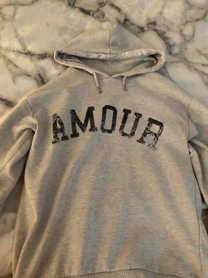 Grå hoodie med tryck 'AMOUR' - Snygg ljusgrå hoodie med huva. På framsidan står det ” amour ” och på ryggen är det en stjärna.❤️ den i ganska bra skick men den är inte lika fluffig inuti 💌💐❤️