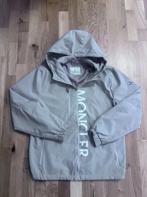 Grå vindjacka från Moncler - Snygg beige vindjacka från Moncler med stor vit logga längs dragkedjan. Jackan har huva, två sidofickor med dragkedja och en bröstficka. Elastiska muddar vid ärmsluten och justerbar nederkant. Tillverkad i ett lätt och vattenavvisande material.