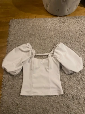 Vit topp med puffärmar från BikBok - Superfin vit topp från BikBok med stora puffärmar och söt detalj med band över axlarna. Toppen har en figurnära passform och är perfekt för dig som vill sticka ut lite extra. Materialet känns mjukt och bekvämt mot huden.