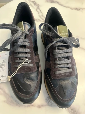 Valentino Garavani camo sneakers - Snygga sneakers från Valentino Garavani med camomönster i svart, grått och mörkblått. Skorna har gråa skosnören, detaljer i mocka och läder samt en klassisk låg siluett. Yttersulan är i gummi med mönstrad undersida för extra grepp.