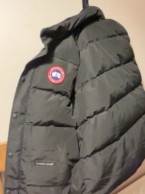 Svart dunjacka från Canada Goose - Säljer en svart dunjacka från Canada Goose med klassisk logotyp på bröstet och diskret varumärkesbrodyr på fickan. Jackan har en hög krage, breda kanaler och är fylld med dun för extra värme. Perfekt för kalla vinterdagar.