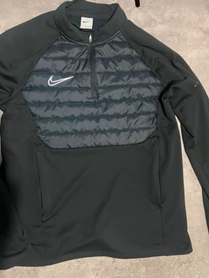 Svart träningsjacka från Nike - Snygg svart träningsjacka från Nike med quiltad frontpanel och halv dragkedja. Jackan har lång ärm, hög krage och vit Nike-logga på bröstet. Perfekt för löpning eller gym. Materialet är en mix av syntet och vadderad front för extra värme.