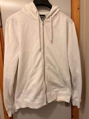 Vit zip-hoodie från J.Lindeberg - Vit hoodie med dragkedja från J.Lindeberg. Klassisk modell med huva, dragsnören och två fickor framtill. Diskret logga broderad på bröstet. Mjuk bomullskänsla och ribbade muddar vid ärmslut och nederkant.