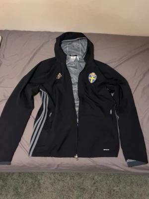 Svart Adidas vindjacka Sverige - Svart vindjacka från Adidas med Sveriges landslagsemblem på bröstet. Jackan har huva, hel dragkedja, två sidofickor med dragkedja och gråa detaljer samt klassiska Adidas-ränder på sidorna. Materialet är lätt och vindtätt, perfekt för sportiga dagar.