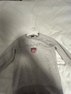 Grå sweatshirt från GANT  - Snygg grå sweatshirt från GANT med broderad logga och sköld med stjärnor och ränder på bröstet. Tröjan har rund halsringning och långa ärmar. Tillverkad i mjukt bomullsmaterial som är skönt mot huden. Perfekt för en avslappnad och stilren look.