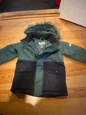 Grön och svart dunjacka från Kaxs - Säljer en grön och svart dunjacka från Kaxs med avtagbar huva och snyggt fuskpäls på kanten. Jackan har stora fickor med knappar framtill och är perfekt för kalla vinterdagar. Materialet är tåligt och håller värmen bra. Passar dig som vill ha en cool och praktisk vinterjacka.