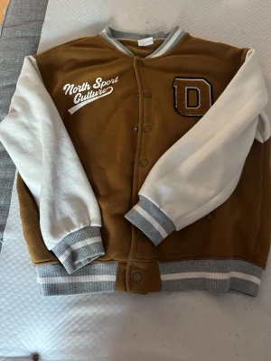 Brun och vit bomberjacka Michigan - Snygg bomberjacka i brun och vit med grå ribbade muddar. Framsidan har tryck med texten 'North Sport Culture' och ett stort D-märke. Baksidan har tryck med 'Michigan Detroit Club'. Jackan har tryckknappar och collegeinspirerad stil.