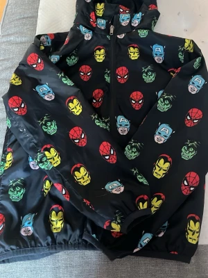 Marvel vår med superhjältar - Cool svart vår jacka med färgglada ansikten av Marvels superhjältar som Spiderman, Iron Man, Hulk och Captain America. Stort Marvel-tryck på ryggen, dragkedja framtill och huva. Perfekt för cosplay eller maskerad.