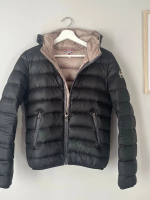 Colmar jacka - Snygg svart dunjacka från Colmar med beige innerfoder och huva. Jackan har två dragkedjeförsedda fickor framtill och quiltad design med breda kanaler. Märkeslogga på ärmen och dragkedja framtill. Perfekt för höst och vår!