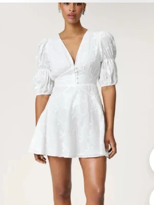 Vit kortklänning med puffärmar - Säljer en vit kortklänning med v-ringning och puffiga kortärmar. Klänningen har markerad midja och subtilt blommigt mönster. Perfekt för dig som vill ha en feminin och trendig look.