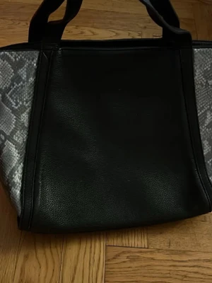 Svart handväska med ormmönster - Snygg svart handväska i konstläder med detaljer i grått ormmönster på sidorna. Väskan har två rejäla handtag och en praktisk ytterficka med dragkedja. Perfekt för dig som gillar en stilren look med lite edge.