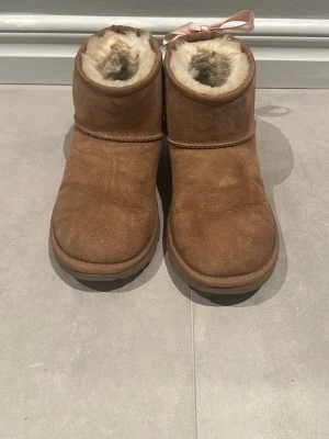 Bruna boots i mocka med fårskinn - Supersköna bruna boots i mocka med fluffigt vitt fårskinnsfoder och en söt ljusrosa rosett bak. Bootsens klassiska form och robusta sula gör dem perfekta för kalla dagar. Materialet är mocka på utsidan och fårskinn på insidan för extra värme. Rosetterna har jag själv satt på.