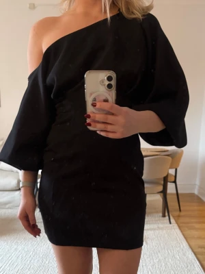 Svart oneshoulder klänning ASOS DESIGN - Säljer en svart oneshoulder klänning från ASOS DESIGN. Klänningen har en kort längd, lös passform i ärmen och markerad midja. Perfekt för dig som gillar en stilren och trendig look med unik ärmdesign.