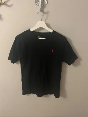 Svart t-shirt från Polo Ralph Lauren - Klassisk svart t-shirt från Polo Ralph Lauren med rund halsringning och den ikoniska röda logotypen broderad på bröstet. Tillverkad i mjuk bomull som känns skön mot huden. Perfekt för en clean och stilren look.