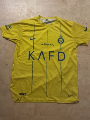 Al Nassr fotbollströja - Snygg gul Al Nassr matchtröja från Nike med blå detaljer och tryck. Framsidan har klubbmärke, sponsorloggor och Nike-logga, medan baksidan har RONALDO och nummer 7 i blått. Tillverkad i lätt och ventilerande material, perfekt för fotbollsträning eller match.