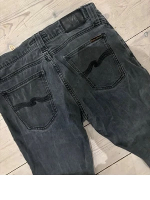 Svarta jeans från Nudie Jeans - Säljer ett par svarta jeans från Nudie Jeans med klassisk femficksmodell och svarta sömmar på bakfickorna. Jeansen har en rak passform och är tillverkade i denim med en tvättad look. Perfekta för en avslappnad stil.
