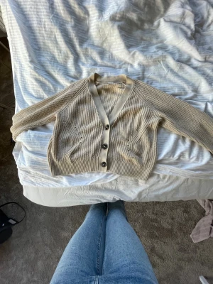 Beige stickad kofta med knappar - Snygg beige stickad kofta med tre knappar framtill. Koftan har ribbade muddar och ett diskret mönster längs sidorna. Croppad modell med lång ärm, perfekt att slänga över en t-shirt eller linne.