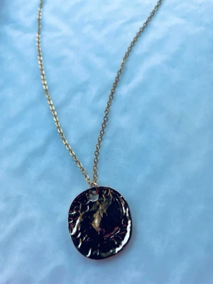 By prisförslag- Guldigt halsband med hamrad rund berlock - Snyggt halsband i guldfärgad metall med en rund, hamrad berlock som ger en cool och trendig look. Kedjan är tunn och smidig, perfekt för att lyfta vilken outfit som helst. Märke och exakt material är okänt.