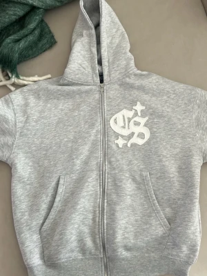 Grå zip hoodie med broderad logga - Säljer en grå zip hoodie  med broderad vit logga och stjärnor på bröstet. Hoodien har huva, två fickor fram och ribbade muddar. Perfekt för chill dagar och streetwear-stil. Den är helt ny använd en gång 