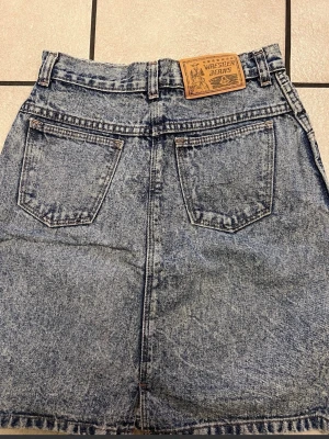 Blå jeanskjol från Wrestler Jeans - Snygg blå jeanskjol från Wrestler Jeans med klassisk femficksdesign och bälteshällor. Kjolen har en rak passform och är tillverkad i slitstark denim med en cool tvättad look. Perfekt för dig som gillar en avslappnad och trendig stil. Wrestler Jeans blå jeans kjol storlek S, midja 66 cm, 100% bomull