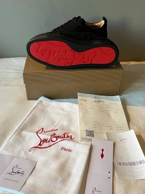 Svarta skor Louboutin  - Svarta sneakers från Louboutin med ikonisk röd sula och nitar framtill. Skorna har snörning och är tillverkade i skinn med en lyxig känsla. Perfekta för dig som vill sticka ut med en edgy och exklusiv look. 